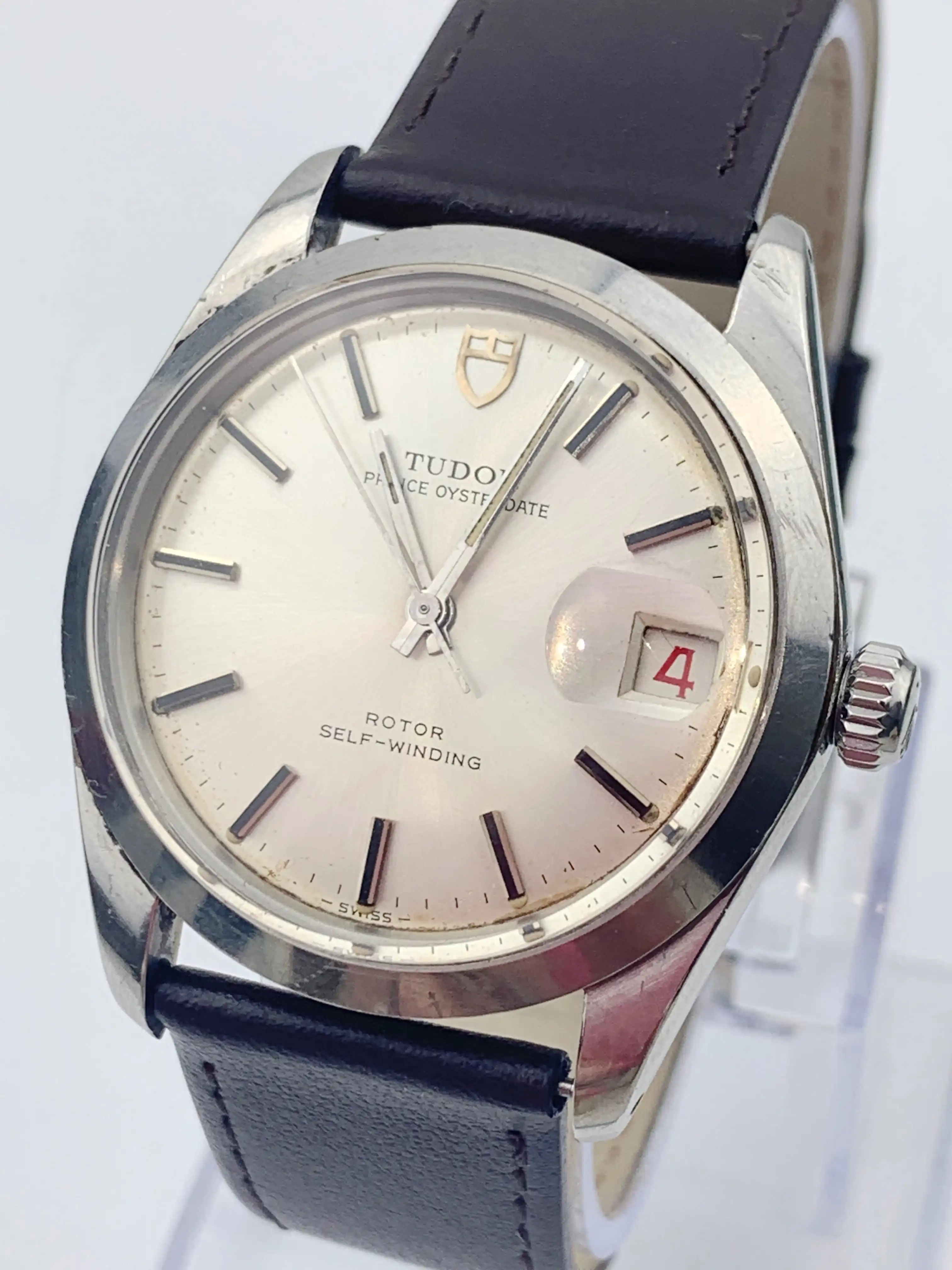 Close-up of a Tudor wristwatch with a silver dial, baton hour markers, and a black leather strap showing the date '4' in a magnified window